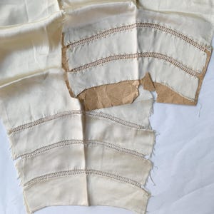 May include: Cream-coloured fabric pieces with decorative stitching. The fabric has a smooth, slightly shiny texture, possibly silk. The pieces are cut into curved shapes, potentially for a garment or craft project. Some pieces are on brown paper.