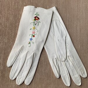 May include: A pair of white leather gloves with colorful floral embroidery. The embroidery features red, yellow, pink, and blue flowers with green stems. The gloves are laid out on a textured, neutral-toned surface.