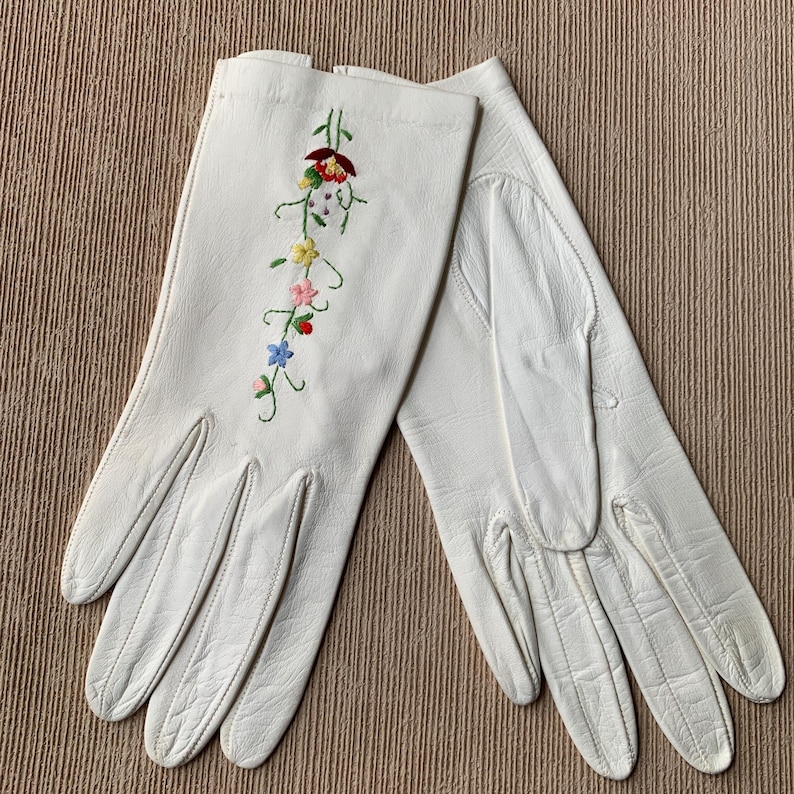 May include: A pair of white leather gloves with colorful floral embroidery. The embroidery features red, yellow, pink, and blue flowers with green stems. The gloves are laid out on a textured, neutral-toned surface.
