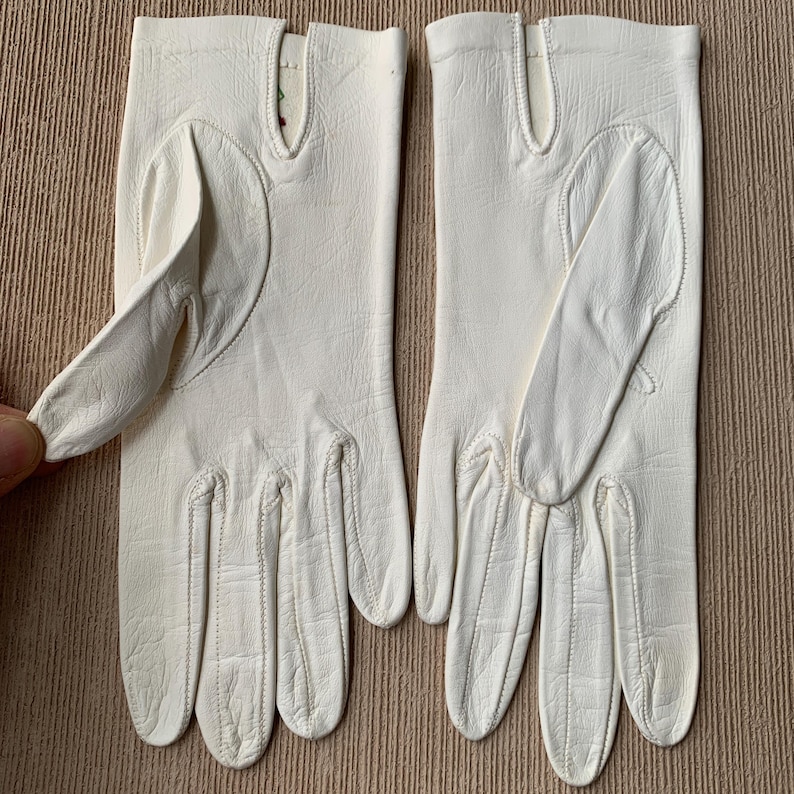 May include: A pair of off-white leather gloves. The gloves have a classic design with a smooth texture and visible stitching. The fingers are neatly shaped, and the gloves appear to be in good condition.