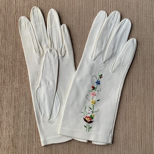 May include: A pair of off-white leather gloves. One glove features a floral embroidery design with red, blue, pink, and yellow flowers. The gloves are laid out on a neutral-toned surface.