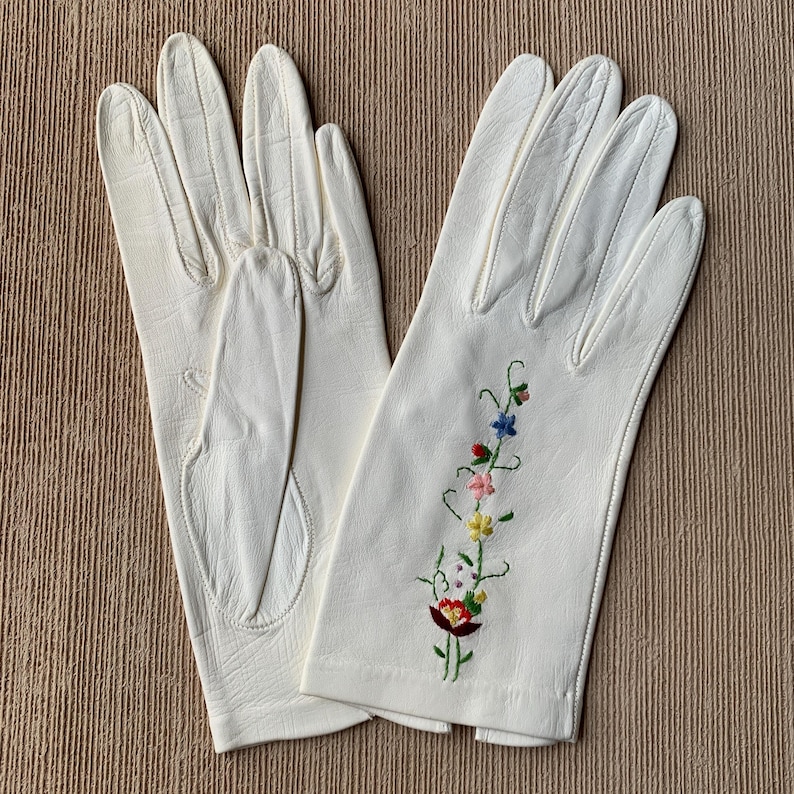 May include: A pair of off-white leather gloves. One glove features a floral embroidery design with red, blue, pink, and yellow flowers. The gloves are laid out on a neutral-toned surface.