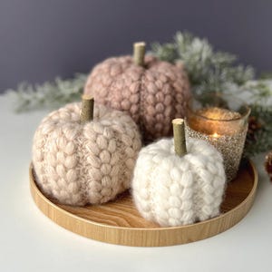 May include: Three round, hand-knitted pumpkins in shades of dusty rose, beige, and cream, each topped with a small wooden stem. The pumpkins are arranged on a circular wooden tray alongside a glass candle holder containing a lit tea light.