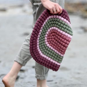 May include: A handmade, half-circle rug with a textured, bobble stitch design. The rug features concentric stripes of burgundy, green, and pink. The rug is held by a person wearing grey trousers.