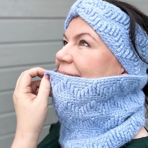 Puede incluir: Un conjunto de diadema y cuello de punto de ganchillo azul claro. La diadema se lleva en la cabeza y el cuello se lleva alrededor del cuello. Ambas piezas están hechas con un punto texturizado.