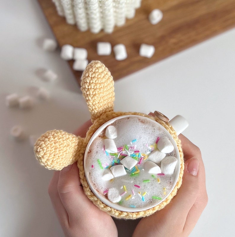 May include: A crocheted bunny holding a white mug filled with hot chocolate and marshmallows topped with colorful sprinkles.