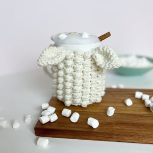 May include: White crocheted mug cozy with a white mug filled with a hot beverage and marshmallows. A cinnamon stick is sticking out of the mug. There are white marshmallows scattered on a wooden surface.