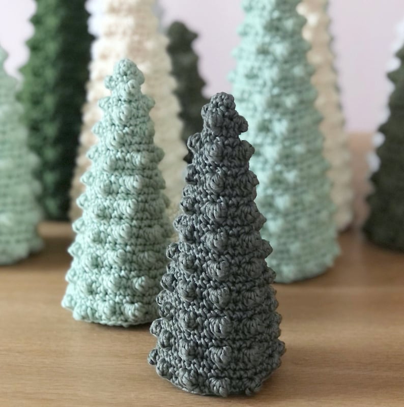 K&ouml;nnte beinhalten: Eine Sammlung geh&auml;kelter Weihnachtsb&auml;ume in verschiedenen Gr&uuml;nt&ouml;nen und Wei&szlig;. Die B&auml;ume sind in einer Reihe auf einer Holzoberfl&auml;che angeordnet.