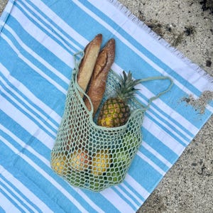 Puede incluir: Una bolsa de red de ganchillo verde claro llena de pi&ntilde;a, naranjas, limas y dos baguettes. La bolsa est&aacute; sobre una toalla de playa a rayas azules y blancas, ideal para un picnic.