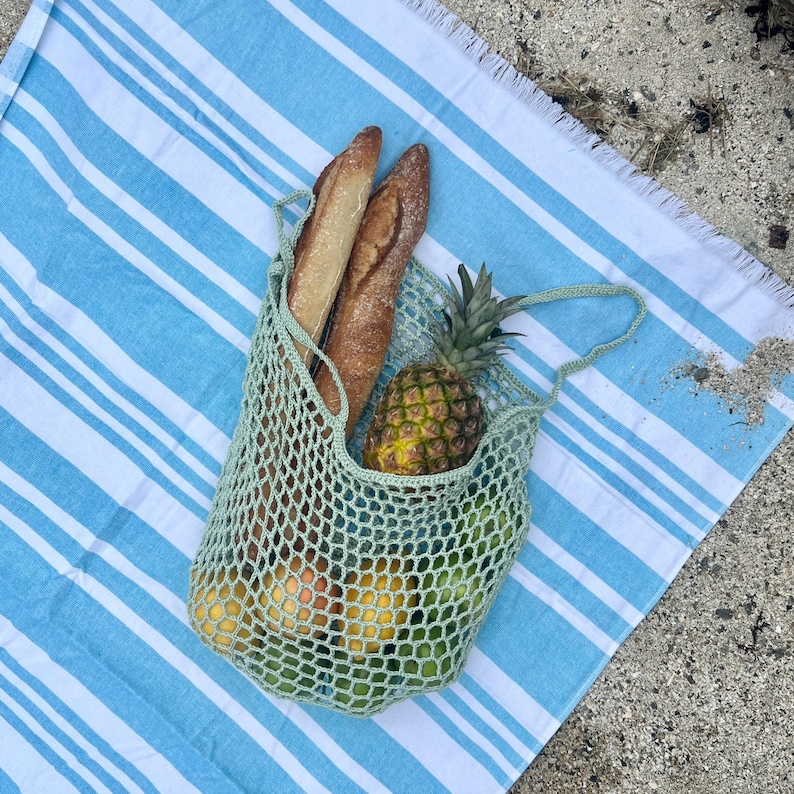 Puede incluir: Una bolsa de red de ganchillo verde claro llena de pi&ntilde;a, naranjas, limas y dos baguettes. La bolsa est&aacute; sobre una toalla de playa a rayas azules y blancas, ideal para un picnic.
