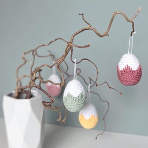 Könnte beinhalten: Vier gehäkelte Ostereier hängen an einem Zweig. Die Eier sind weiß mit verschiedenen farbigen Streifen. Die Eier sind grün, rosa, gelb und weiß.