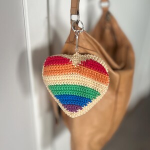 Puede incluir: Un llavero en forma de coraz&oacute;n tejido a ganchillo con los colores del arco&iacute;ris: rojo, naranja, amarillo, verde, azul y morado. El llavero est&aacute; sujeto a un llavero plateado y cuelga delante de un bolso de cuero marr&oacute;n.