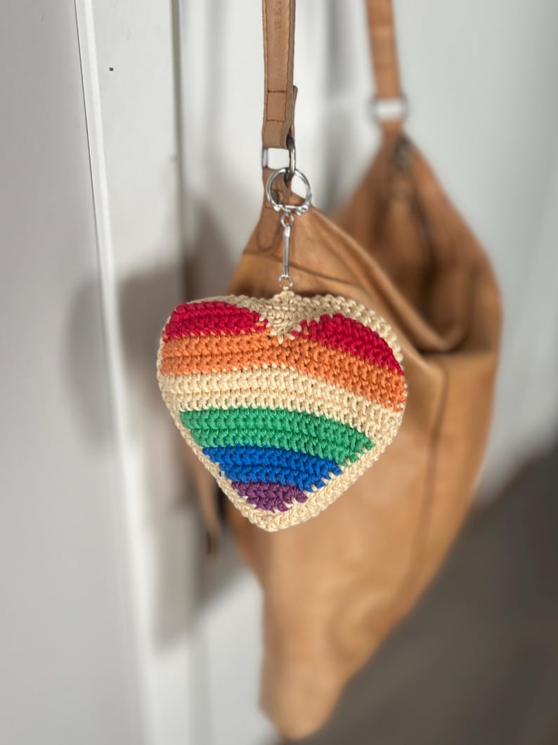 Puede incluir: Un llavero en forma de coraz&oacute;n tejido a ganchillo con los colores del arco&iacute;ris: rojo, naranja, amarillo, verde, azul y morado. El llavero est&aacute; sujeto a un llavero plateado y cuelga delante de un bolso de cuero marr&oacute;n.