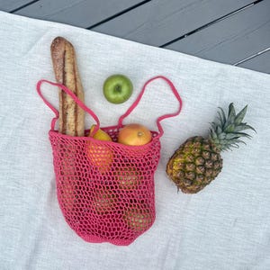Puede incluir: Una bolsa de mano de ganchillo rosa brillante llena de productos frescos, incluyendo una baguette, manzanas y naranjas. Una pi&ntilde;a entera y una manzana verde est&aacute;n junto a la bolsa, todo sobre un pa&ntilde;o blanco.