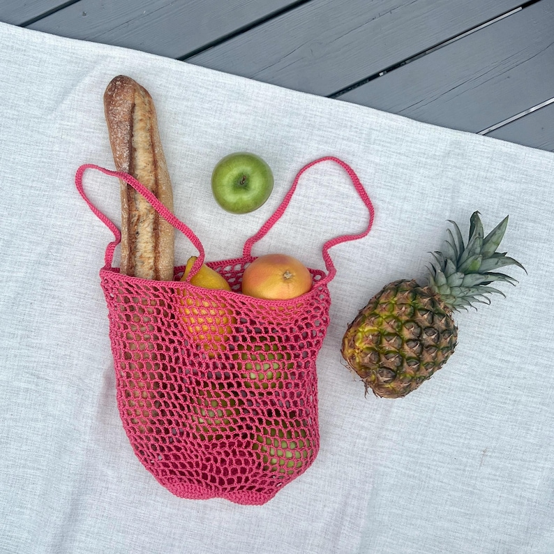 Puede incluir: Una bolsa de mano de ganchillo rosa brillante llena de productos frescos, incluyendo una baguette, manzanas y naranjas. Una pi&ntilde;a entera y una manzana verde est&aacute;n junto a la bolsa, todo sobre un pa&ntilde;o blanco.