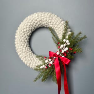 May include: A cream-coloured, knitted wreath with a festive design. The wreath features evergreen sprigs, white berries, and red berries, and is accented by a vibrant red satin bow. A seasonal decoration.
