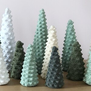 May include: A collection of crocheted Christmas trees in various shades of green, white, and gray. The trees are all different sizes and have a textured, bumpy surface.