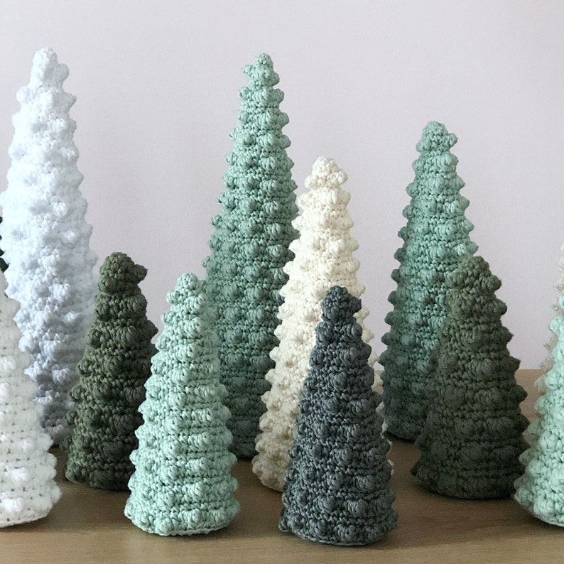 May include: A collection of crocheted Christmas trees in various shades of green, white, and gray. The trees are all different sizes and have a textured, bumpy surface.