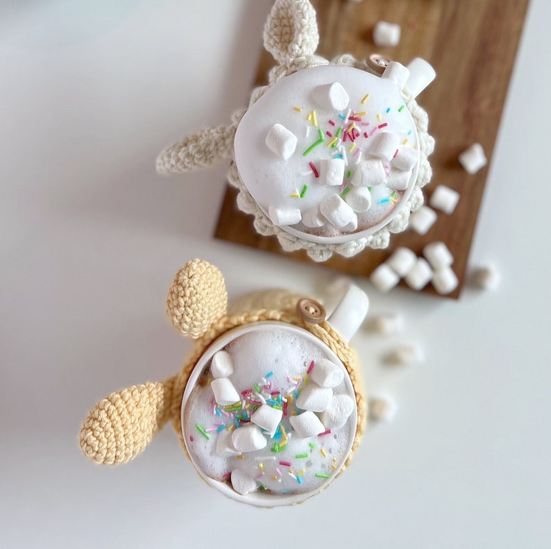 May include: Two crocheted mugs with handles shaped like animal ears. Both mugs are filled with hot chocolate topped with marshmallows and colorful sprinkles.