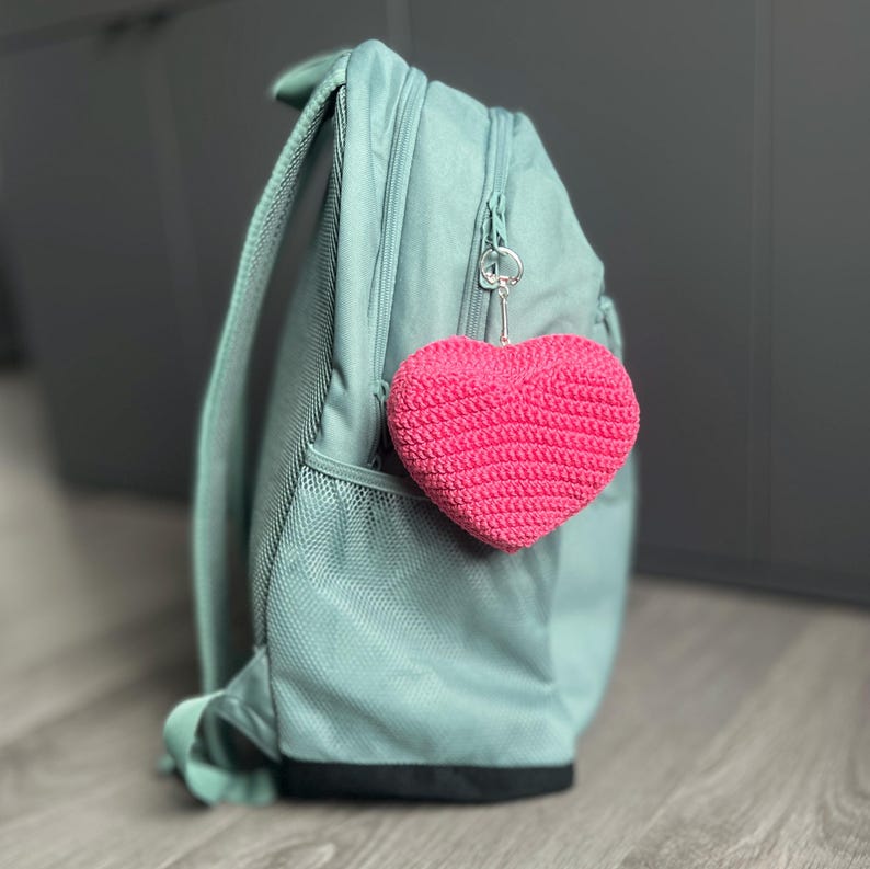 Puede incluir: Un llavero en forma de coraz&oacute;n rosa brillante, tejido a ganchillo, colgado de una mochila azul claro. El coraz&oacute;n est&aacute; hecho de hilo texturizado y est&aacute; unido a un llavero plateado. La mochila tiene un bolsillo con cremallera y un bolsillo lateral de malla.