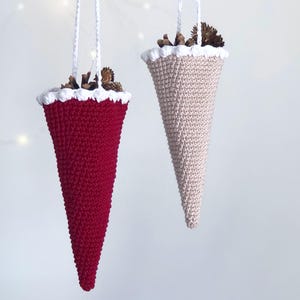 May include: Two crocheted cone-shaped Christmas ornaments hanging from white string. The ornament on the left is red with a white trim. The ornament on the right is beige with a white trim. Both ornaments are filled with small pine cones.