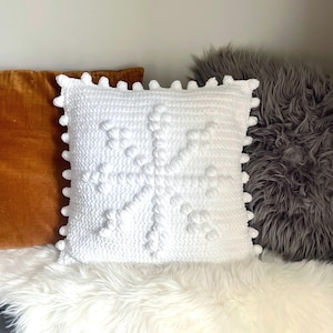 May include: A square white crochet pillow with a snowflake design. The pillow has a textured surface with raised bobbles and a pom-pom trim around the edges. It is placed on a white faux fur surface, with other pillows in the background.
