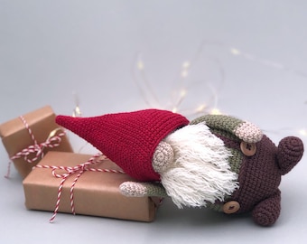 May include: A crocheted gnome with a red hat, brown body, green pants, and a white beard. The gnome is lying on its side next to two wrapped gifts.