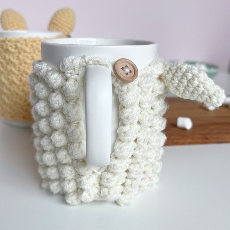 May include: White crocheted mug cozy with a wooden button. The cozy is shaped like a sheep with a small ear sticking out.