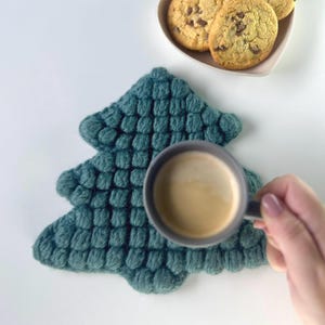 Puede incluir: Un posavasos en forma de árbol de Navidad, texturizado y de color verde azulado. Una taza gris llena de café se sostiene sobre el posavasos. Un plato pequeño con galletas con chispas de chocolate está en el fondo.