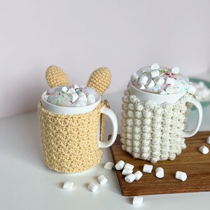 May include: Two white mugs with crocheted covers, one yellow and one white, filled with hot chocolate and marshmallows. The mugs are on a wooden surface with white marshmallows scattered around.