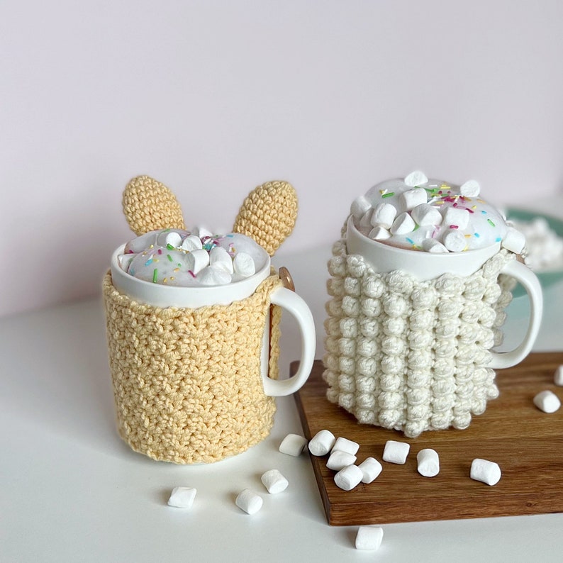 May include: Two white mugs with crocheted covers, one yellow and one white, filled with hot chocolate and marshmallows. The mugs are on a wooden surface with white marshmallows scattered around.
