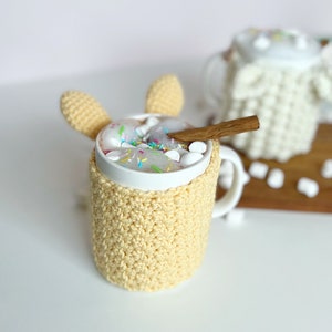 May include: A white mug with a crocheted yellow cozy and bunny ear handles. The mug is filled with hot chocolate topped with marshmallows and colorful sprinkles. A cinnamon stick rests on the rim of the mug.