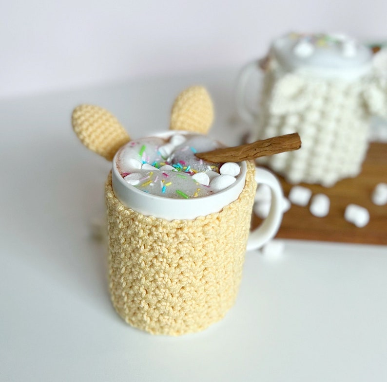 May include: A white mug with a crocheted yellow cozy and bunny ear handles. The mug is filled with hot chocolate topped with marshmallows and colorful sprinkles. A cinnamon stick rests on the rim of the mug.
