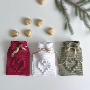 May include: Three crocheted gift bags in red, white, and olive green, each with a heart design and tied with a gold ribbon. Gold heart-shaped candies are scattered around the bags. A sprig of evergreen adds a festive touch.