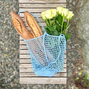 Puede incluir: Una bolsa de red de ganchillo azul claro contiene dos baguettes y un ramo de rosas amarillas. La bolsa cuelga de un banco de madera. El tejido abierto de la bolsa es visible, y las baguettes tienen una corteza dorada.