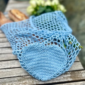 Puede incluir: Una bolsa de ganchillo azul claro con un detalle en forma de coraz&oacute;n. La bolsa tiene un dise&ntilde;o de red y est&aacute; sobre una superficie de madera. La bolsa est&aacute; llena de una baguette y algunos tallos verdes.