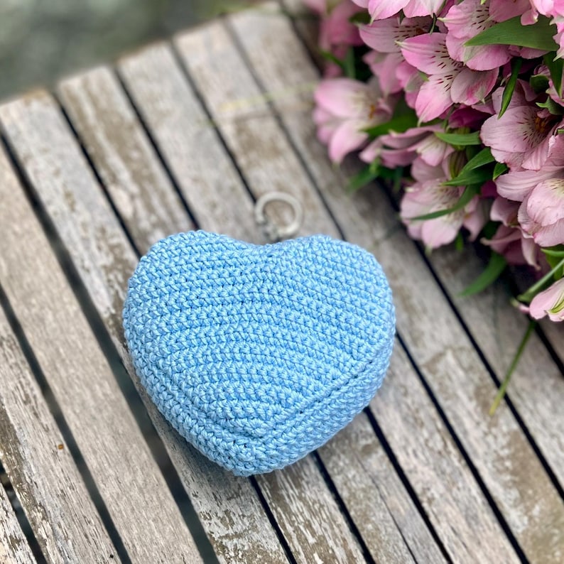 Puede incluir: Un objeto de ganchillo en forma de coraz&oacute;n azul claro con un llavero plateado. El art&iacute;culo hecho a mano se coloca sobre una superficie de madera desgastada, con flores rosas en el fondo. El art&iacute;culo de ganchillo es de un material suave y texturizado.