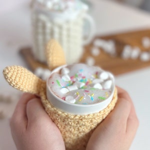 May include: A white mug with a crocheted cozy in a light yellow color. The mug is filled with hot chocolate topped with marshmallows and colorful sprinkles.