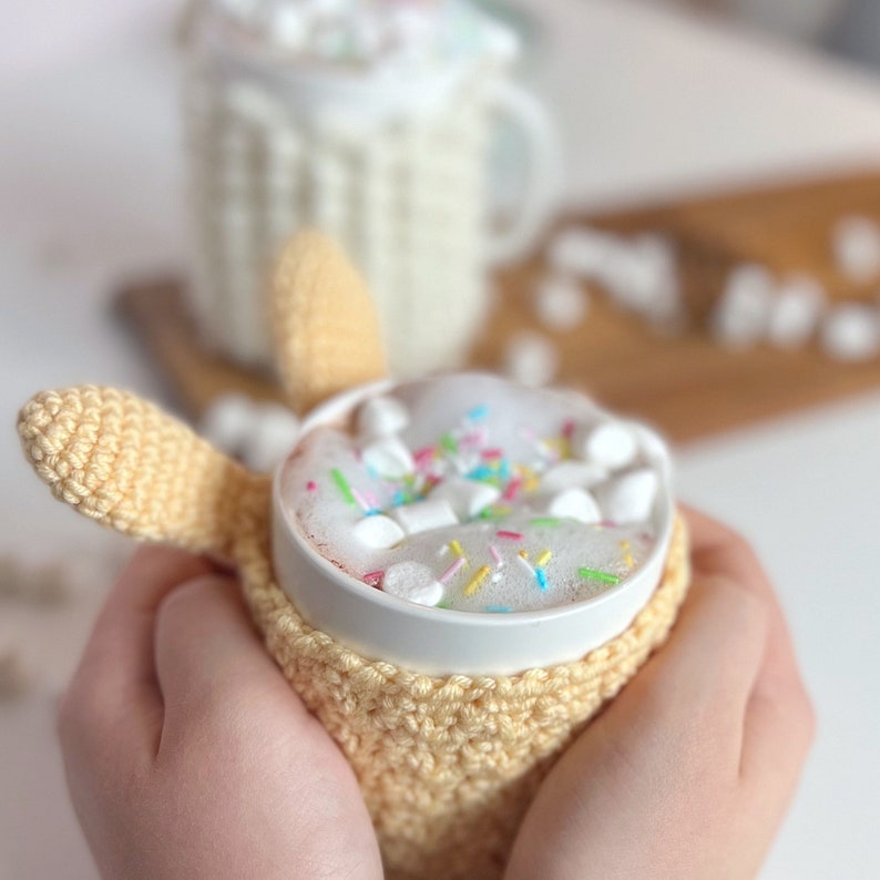 May include: A white mug with a crocheted cozy in a light yellow color. The mug is filled with hot chocolate topped with marshmallows and colorful sprinkles.