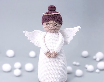 May include: A white crocheted angel doll with brown hair and a gold halo. The angel is standing on a white surface with white balls scattered around it.