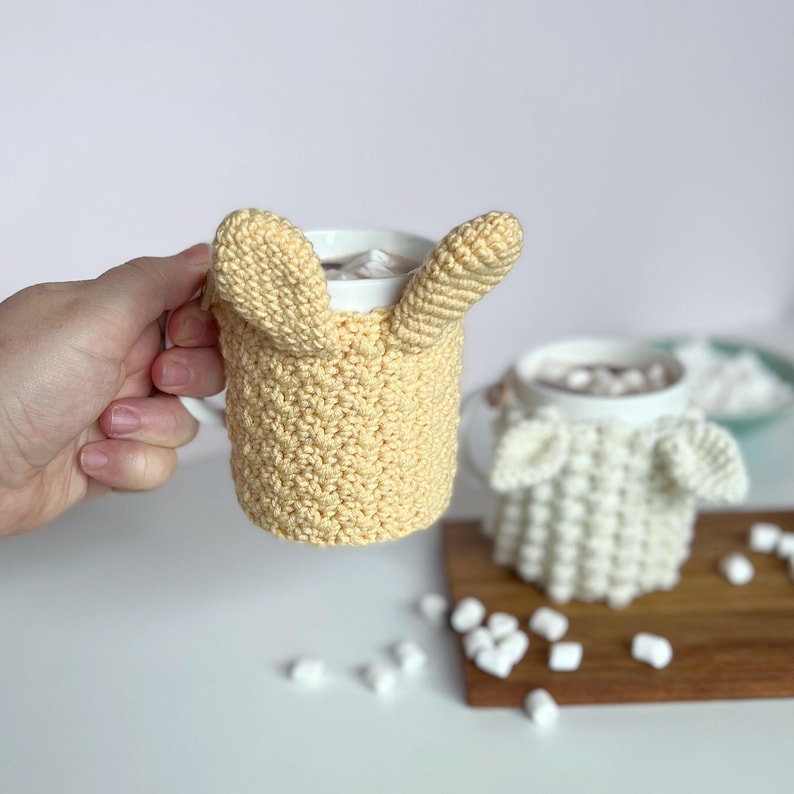 May include: Two crocheted mug cozies in the shape of animal faces. The yellow cozy has bunny ears and the white cozy has sheep ears. Both cozies are on mugs filled with hot chocolate and marshmallows.