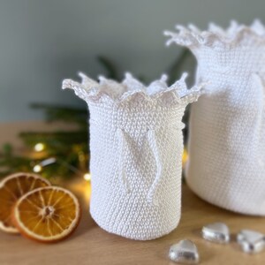 May include: Two white crocheted bags with a scalloped edge and a drawstring closure. The bags are sitting on a wooden surface with dried orange slices and silver heart-shaped decorations.