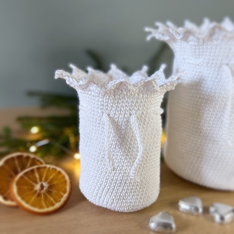 May include: Two white crocheted bags with a scalloped edge and a drawstring closure. The bags are sitting on a wooden surface with dried orange slices and silver heart-shaped decorations.