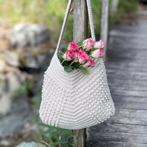 May include: A white crocheted tote bag with a textured design, hanging on a wooden fence post. The bag is filled with pink roses.