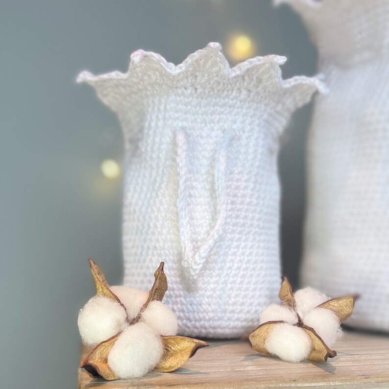 May include: Two white crocheted bags with a ruffled top and a drawstring closure. The bags are sitting on a wooden surface with cotton bolls.