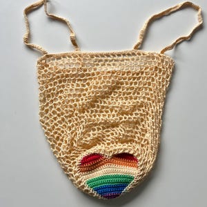 Puede incluir: Un bolso tote beige tejido a ganchillo con un dise&ntilde;o de coraz&oacute;n arco&iacute;ris. El bolso presenta un tejido similar a una red y dos correas beige. El coraz&oacute;n arco&iacute;ris est&aacute; hecho de hilo rojo, naranja, amarillo, verde, azul y morado.