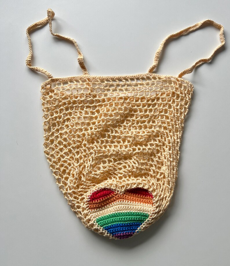 Puede incluir: Un bolso tote beige tejido a ganchillo con un dise&ntilde;o de coraz&oacute;n arco&iacute;ris. El bolso presenta un tejido similar a una red y dos correas beige. El coraz&oacute;n arco&iacute;ris est&aacute; hecho de hilo rojo, naranja, amarillo, verde, azul y morado.