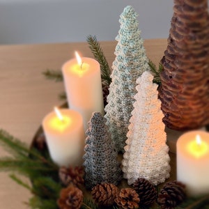 K&ouml;nnte beinhalten: Drei geh&auml;kelte Weihnachtsb&auml;ume in Wei&szlig;, Hellblau und Grau, mit drei brennenden Kerzen davor. Die B&auml;ume stehen auf einem Holztisch mit Tannenzweigen und Tannenzapfen drumherum.