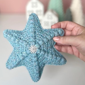 以下が含まれることがあります： 水色の星型かぎ針編みオーナメント。表面は質感があり、中央には銀色の雪の結晶が飾られています。手作りの装飾品で、お祭りや季節の飾り付けに適しています。サイズは約15cmです。