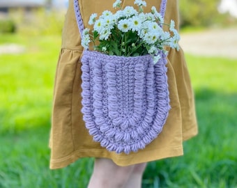Pode incluir: Uma bolsa de crochê lavanda com um design texturizado e volumoso. A bolsa é usada como bolsa transversal e está cheia de um buquê de margaridas brancas.